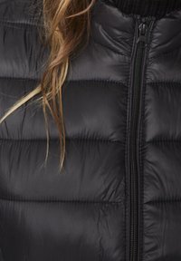 Černá prošívaná bunda se zipem vpředu a několika prameny světle hnědých vlasů spadajícími přes levé rameno.