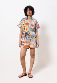 Kurzärmliger, kragenloser Jumpsuit im mehrfarbigen Print mit verschiedenen Mustern. Verfügt über eine taillierte, mit Gürtel versehenen Taille und lebhafte, tropische Motive. Rote Sandalen.