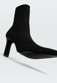 Bottine noire à tige tricotée ajustée et à talon haut et carré. La semelle est lisse, avec des accents texturés subtils.