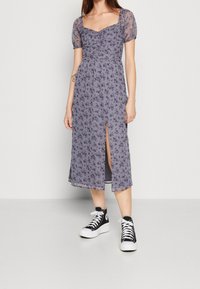 Šaty s květinovým potiskem v světle fialové barvě, s nabíranými rukávy, čtvercovým výstřihem a bočním rozparkem; nošeny s černými vysokými teniskami.