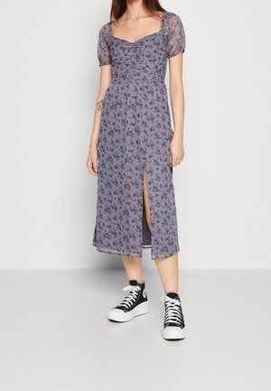 Blomstertrykt kjole i lys lilla med puffærmer, firkantet halsudskæring og sideslids; båret med sorte højskårne sneakers.