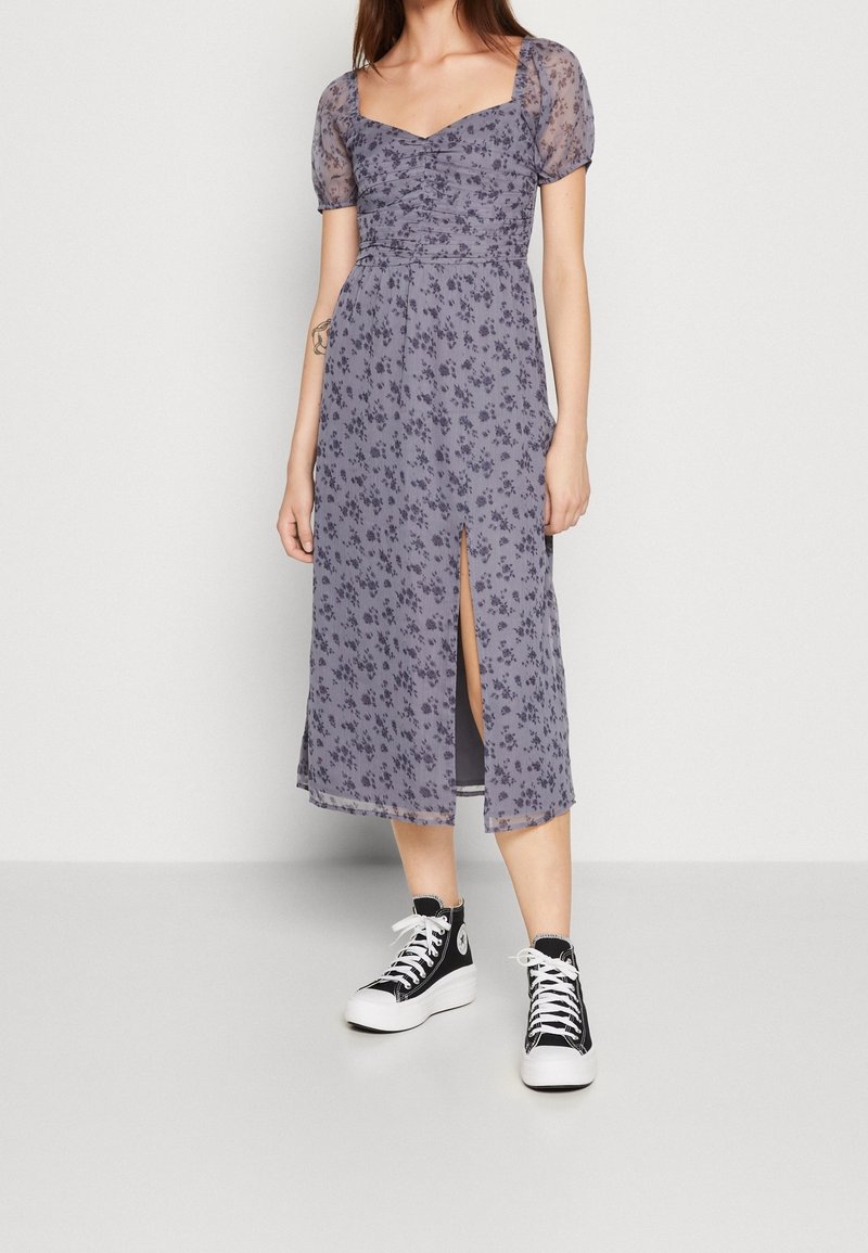 Šaty s květinovým potiskem v světle fialové barvě, s nabíranými rukávy, čtvercovým výstřihem a bočním rozparkem; nošeny s černými vysokými teniskami.