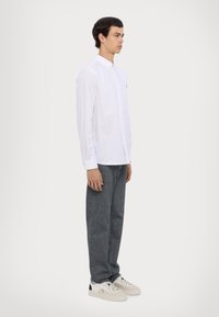 White long-sleeve shirt with buttoned collar, grey straight-leg jeans, and white sneakers with black accents. Model stands in profile against a light background.