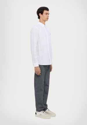 Hvit langermet skjorte med knappelukking på kragen, grå rette jeans og hvite joggesko med svarte detaljer. Modellen står i profil mot en lys bakgrunn.