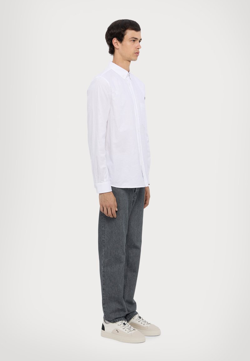 White long-sleeve shirt with buttoned collar, grey straight-leg jeans, and white sneakers with black accents. Model stands in profile against a light background.