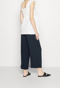 Bílý texturovaný top s volánovými ramínky spárovaný s volnými tmavě modrými širokými kalhotami a černými nazouvacími botami.