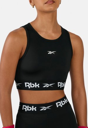 Donna che indossa un reggiseno sportivo Reebok nero e leggings con bande bianche con logo, mostrata dalle spalle alla vita su uno sfondo uniforme.