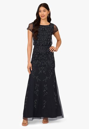 Donna che indossa un abito nero lungo fino a terra con ricamo floreale, maniche corte trasparenti e tacchi aperti color nudo, in piedi con una mano sul fianco.