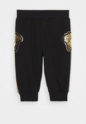 Pantalon noir court avec poignets et taille côtelés, présentant des logos tête de Méduse dorés de chaque côté près des hanches et des bandes latérales dorées.