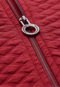 Tela acolchada roja con un tirador de cremallera de metal plateado y una etiqueta circular que dice "StreetOne". El patrón texturizado añade interés visual.