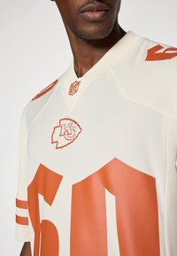 Valkoinen jalkapallopaita oransseilla yksityiskohdilla, jossa on suuri numero 60, joukkueen logo ja NFL-embleemi. Rypytetty pinta ja V-muotoinen kaula-aukko.