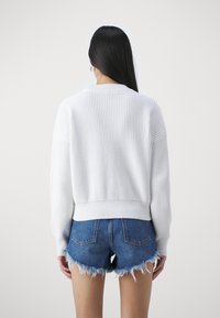 Maglione bianco lavorato a maglia con texture a coste e spalle scese, abbinato a pantaloni corti di denim con orlo sfrangiato.