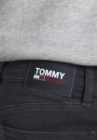 Černé džínové kalhoty s koženou nášivkou. Nálepka obsahuje nápis "TOMMY JEANS" bílým a červeným písmem, spolu s proužkovanými akcenty v červené a bílé barvě.