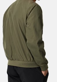 Homme portant une veste bomber vert olive avec une taille élastique et un pantalon noir, main dans la poche, sur un fond uni.