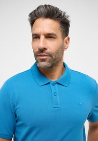 Tyrkysové polo tričko z texturované látky, s rozepínacím límcem, dvouknoflíkovou légou a vyšitým logem na levé hrudi.