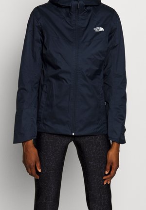 Osoba nosząca czarną kurtkę na zamek błyskawiczny z logo The North Face na piersi i ciemne legginsy z wzorem.