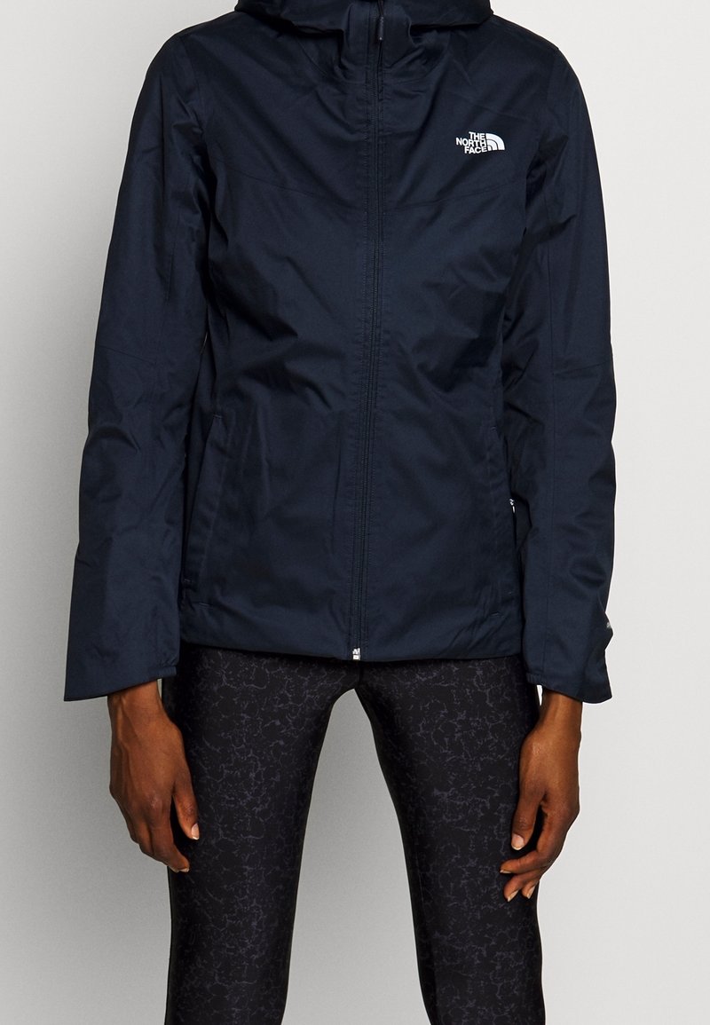 Personne portant une veste noire zippée avec le logo The North Face sur la poitrine et des leggings foncés à motifs.
