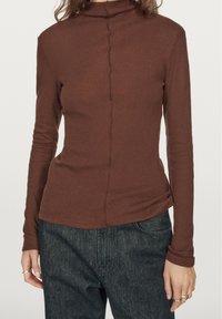 Braunes Langarmoberteil mit hohem Kragen, aus weichem Rippmaterial gefertigt. Mit einer Naht in der Mitte und taillierter Silhouette. Kombiniert mit dunklen Jeans.