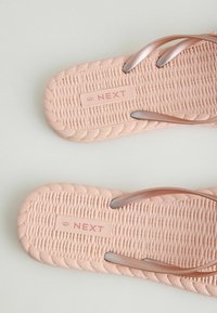 Dvivietė smėlio spalvos pintų šlepečių pora su metalinėmis juostelėmis, ant įdėklų pažymėta „NEXT“ etiketė ir 6 dydis, ant šviesaus fono.