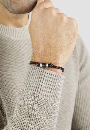 Bracelet en corde noire avec des accents argentés, au design texturé, porté au poignet, complété par un pull gris clair.