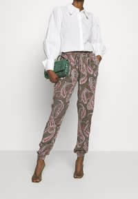 Femme portant un blouse blanche à manches bouffantes, un pantalon à motifs paisley, des sandales à talons vertes et tenant un sac à main texturé vert.