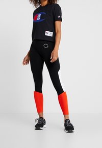 Černé zkrácené tričko s modrým a červeným grafickým logem; v kombinaci s černými legínami s bílým a oranžovým barevným blokem. Černé sportovní boty.