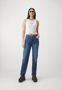 Top corto sin mangas blanco, pantalones vaqueros rectos azules, zuecos negros, gafas de sol rosa estilo cat-eye, collares en capas, de pie contra un fondo simple.