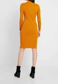 Fasonowany, prążkowany sukienka w intensywnym pomarańczowym kolorze z długimi rękawami i wysokim kołnierzem; posiada guziki na ramionach. Połączona z czarnymi, masywnymi botkami na obcasie.