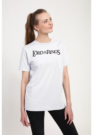 Kvinde med langt hår i hestehale iført hvid t-shirt med sort tekst "The Lord of the Rings", stående mod en ensfarvet baggrund.