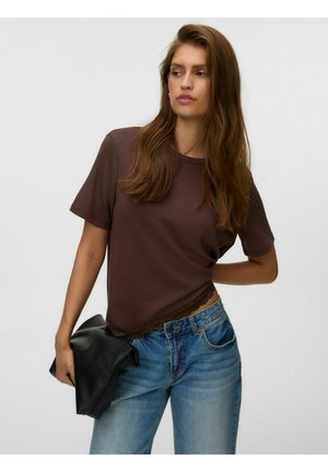 Femme aux longs cheveux bruns porte un haut marron à manches courtes et un jean bleu, tenant une pochette noire d'une main derrière son dos.