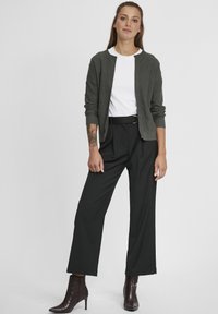 Cardigan texturé vert foncé sur une chemise blanche, associé à un pantalon noir taille haute et des bottines bordeaux. Design simple et neutre.