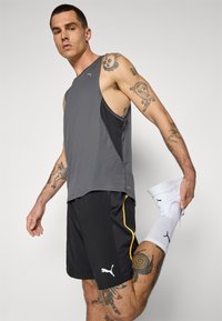 Grå ærmeløs træningstop med sorte accenter, parret med sorte shorts med en gul sidebjælke og hvid Puma-logotype, hvide sneakers.