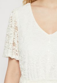 Biela čipkovaná šaty s V-výstrihom, krátkymi, voľnými rukávmi a gombíkovou výzdobou vpredu. Textúra je zložitá s kvetinovými vzormi.