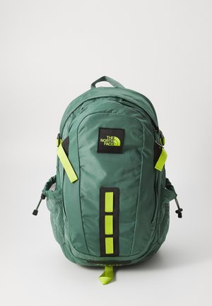 Sac à dos vert North Face avec plusieurs compartiments, tirettes de fermeture éclair jaune vif, poches latérales en mesh et une poignée de transport sur le dessus.