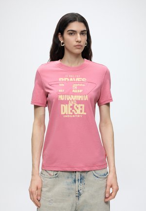 Femeie purtând un tricou roz cu logo și text Diesel galben, asortat cu blugi acid-wash albastru deschis, stând pe un fundal alb simplu.