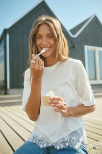 Femme assise en plein air sur une terrasse en bois, souriante en mangeant une glace dans une petite coupe avec une cuillère, portant une chemise blanche et un jean bleu.