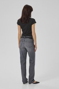 Keskmise pruunide juustega naine, kes kannab tihedalt sobivat musta lühikeste varrukatega topi, hallid sirge säärega teksapükse musta vööga ja musti teravnurkseid baleriinajalatsid.