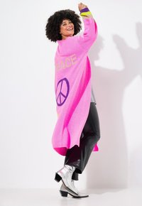 Rosa gestrickte Strickjacke mit dem Schriftzug "PEACE" und einem grafischen Friedenzeichen. Kombiniert mit schwarzen, eng anliegenden Hosen und silbernen hohen Stiefeln. Starke Kontrastfarben.