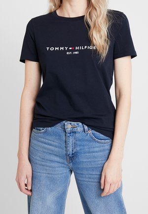 Kobieta ubrana w czarny T-shirt Tommy Hilfiger z logo i napisem "EST. 1985", w połączeniu z wysokim stanem niebieskich dżinsów na jednolitym tle.