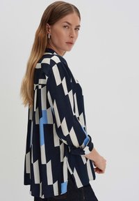 Blusa navy con un motivo geometrico in crema e azzurro chiaro, con maniche lunghe, vestibilità rilassata e polsini arrotolati. Tessuto morbido.