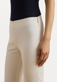 Crème-kleurige broek met een slim-fit ontwerp, voorzien van een zijrits en een gladde, zachte textuur. Hand rustend op het heupgebied.