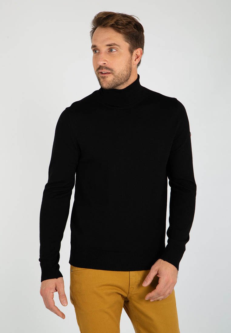 Maglione a collo alto nero con polsini e orlo a costine, indossato con pantaloni color senape. Il tessuto appare liscio e aderente.