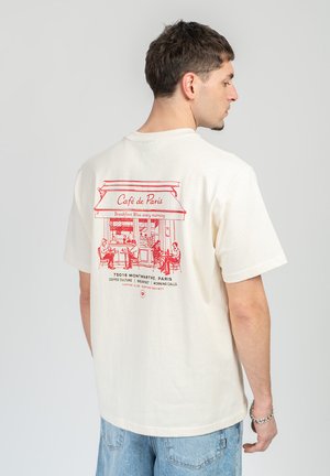 Man draagt een roomkleurig T-shirt met een rood lijntekening van de gevel van Café de Paris en mensen die buiten zitten, met de rug naar de camera toe.