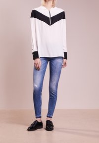 Top de manga larga con un cuerpo blanco y diseño en chevron negro, con cremallera y puños abotonados, combinado con jeans ajustados de color azul claro y zapatos negros.