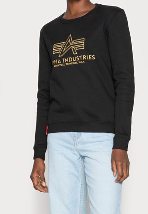 Personne portant un sweat-shirt noir avec un logo Alpha Industries jaune et un jean bleu clair, debout devant un fond uni.