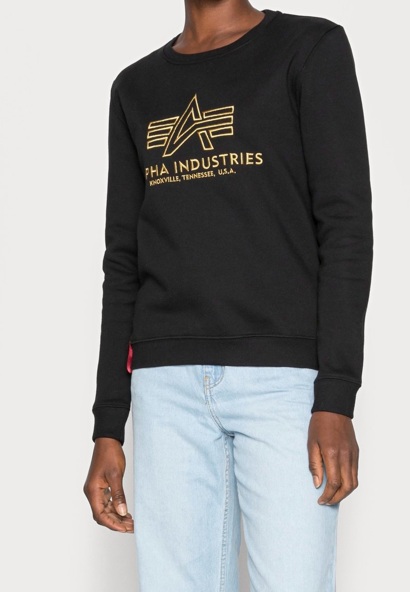 Personne portant un sweat-shirt noir avec un logo Alpha Industries jaune et un jean bleu clair, debout devant un fond uni.