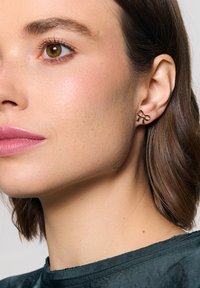 Złote kolczyki w kształcie kokardek, z gładkim wykończeniem i delikatną teksturą, noszone przez osobę z krótkimi włosami.