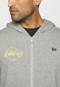 Sudadera gris con cremallera hecha de tela suave, que presenta un logo amarillo de "LOS ANGELES LAKERS" y un emblema negro de New Era en el pecho.