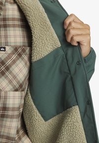 Veste verte avec un doublure beige et duveteuse au col. La chemise à carreaux en dessous présente des motifs bruns et verts. Inclut des détails de boutons.