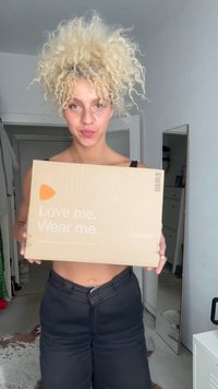 Scatola di cartone marrone con logo arancione e testo bianco "Amami. Indossami." tenuta da una persona, ambientata in un contesto interno neutro.